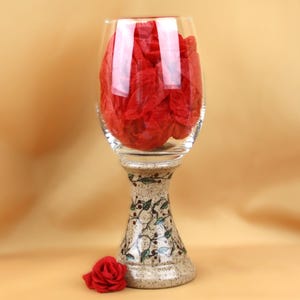May include: A clear wine glass filled with red tissue paper sits atop a decorative pedestal base. The base is beige with a floral design. A red rose rests on the surface next to the base.