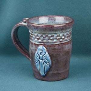 K&ouml;nnte beinhalten: Ein Keramikbecher mit gesprenkelter violetter und brauner Glasur, der ein strukturiertes Band nahe dem Rand und ein erhabenes, hellblaues, blattf&ouml;rmiges Emblem auf der Vorderseite aufweist. Der Becher hat einen stabilen Henkel und eine glatte Oberfl&auml;che.