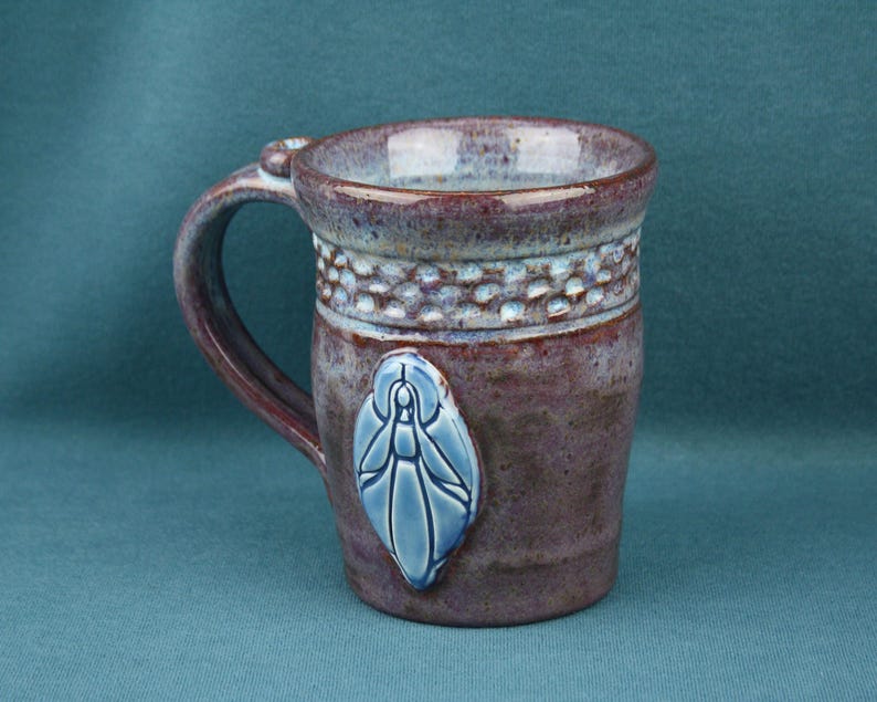 K&ouml;nnte beinhalten: Ein Keramikbecher mit gesprenkelter violetter und brauner Glasur, der ein strukturiertes Band nahe dem Rand und ein erhabenes, hellblaues, blattf&ouml;rmiges Emblem auf der Vorderseite aufweist. Der Becher hat einen stabilen Henkel und eine glatte Oberfl&auml;che.