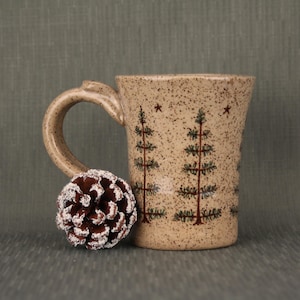 May include: A speckled beige ceramic mug with a curved handle, featuring a hand-painted design of dark green pine trees and small stars. A single brown pinecone with white-tipped scales rests against the base of the mug handle.
