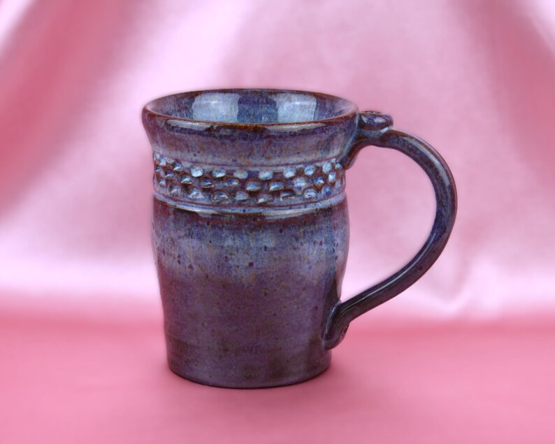 K&ouml;nnte beinhalten: Ein handgefertigter Keramikbecher mit einer tiefvioletten und blauen, gesprenkelten Glasur. Der obere Teil des Bechers zeigt ein strukturiertes Band aus kleinen, erhabenen Vertiefungen um den Rand, erg&auml;nzt durch einen stabilen, geschwungenen Henkel.