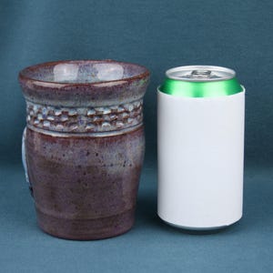 K&ouml;nnte beinhalten: Ein Keramikbecher mit gesprenkelter violetter und brauner Glasur und einem strukturierten Band aus kleinen Vertiefungen nahe dem Rand, platziert neben einer wei&szlig;en Standard-Getr&auml;nkedose mit gr&uuml;nem Deckel zum visuellen Gr&ouml;&szlig;envergleich der Objekte.