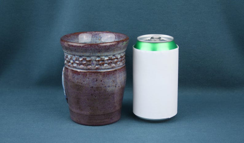 K&ouml;nnte beinhalten: Ein Keramikbecher mit gesprenkelter violetter und brauner Glasur und einem strukturierten Band aus kleinen Vertiefungen nahe dem Rand, platziert neben einer wei&szlig;en Standard-Getr&auml;nkedose mit gr&uuml;nem Deckel zum visuellen Gr&ouml;&szlig;envergleich der Objekte.