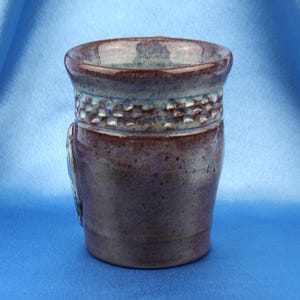 K&ouml;nnte beinhalten: Ein Keramikbecher mit gl&auml;nzender Oberfl&auml;che in tiefen Violett- und Braunt&ouml;nen. Der obere Bereich weist ein strukturiertes Band mit einem sich wiederholenden Quadratmuster auf, w&auml;hrend der Rand in eine hellere blaugraue Glasur &uuml;bergeht.
