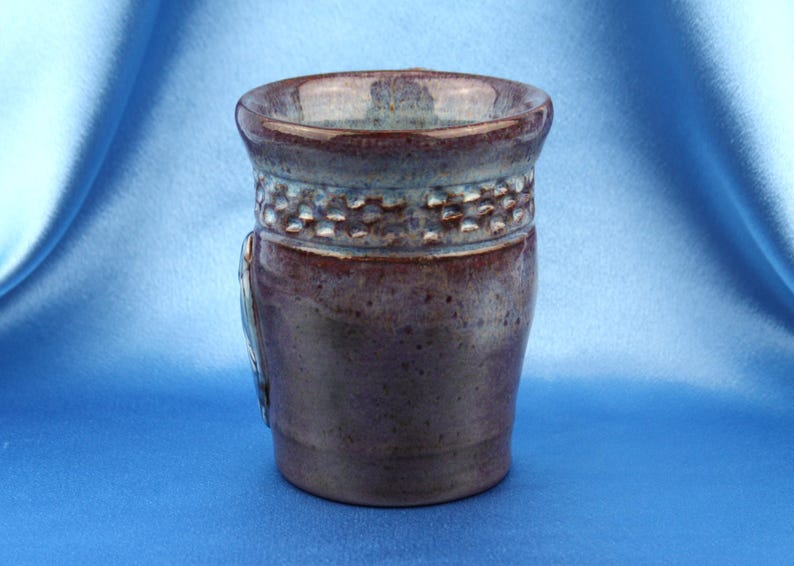 K&ouml;nnte beinhalten: Ein Keramikbecher mit gl&auml;nzender Oberfl&auml;che in tiefen Violett- und Braunt&ouml;nen. Der obere Bereich weist ein strukturiertes Band mit einem sich wiederholenden Quadratmuster auf, w&auml;hrend der Rand in eine hellere blaugraue Glasur &uuml;bergeht.