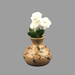 May include: A small, round, speckled beige ceramic vase with a narrow neck, featuring a hand-painted black vine design with green leaves and small red berries. Three artificial white roses with green stems are arranged inside the vase.