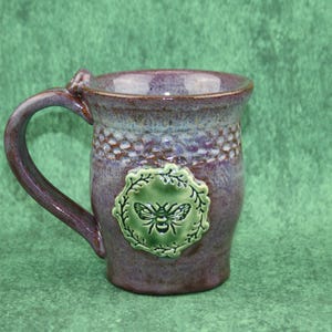 May include: A ceramic mug with a purple and blue glazed finish featuring a textured honeycomb pattern near the rim and a central light green circular medallion embossed with a detailed bee illustration surrounded by a leafy wreath design.
