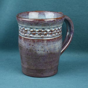 K&ouml;nnte beinhalten: Ein Keramikbecher mit einer gesprenkelten violetten und blauen Glasur und einem texturierten, eingekerbten Band im oberen Bereich. Der Becher hat einen breiten Rand, einen zylindrischen K&ouml;rper, der sich zur Basis hin verj&uuml;ngt, und einen Henkel.