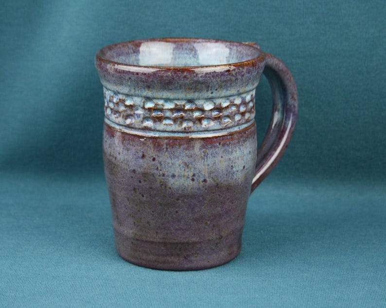 K&ouml;nnte beinhalten: Ein Keramikbecher mit einer gesprenkelten violetten und blauen Glasur und einem texturierten, eingekerbten Band im oberen Bereich. Der Becher hat einen breiten Rand, einen zylindrischen K&ouml;rper, der sich zur Basis hin verj&uuml;ngt, und einen Henkel.