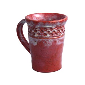 May include: A tall, red ceramic mug with a handle. The mug features a textured band of small, circular indentations near the top, with a white and red speckled glaze. The mug is suitable for hot or cold beverages.