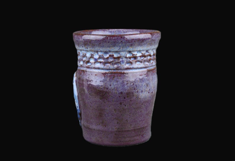 K&ouml;nnte beinhalten: Ein zylindrischer Keramikbecher mit gl&auml;nzender Oberfl&auml;che in Violett- und Blaut&ouml;nen. Der obere Bereich zeigt ein strukturiertes Band mit einem sich wiederholenden eingedr&uuml;ckten Muster, w&auml;hrend der untere Teil eine glatte, gesprenkelte Glasur hat.