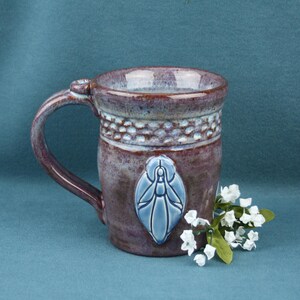 K&ouml;nnte beinhalten: Ein Keramikbecher mit gesprenkelter violetter und blauer Glasur, der ein strukturiertes Band am Rand und ein erhabenes, hellblaues ovales Emblem mit einem stilisierten Insekt aufweist. Ein kleines B&uuml;ndel wei&szlig;er Kunstblumen liegt am Fu&szlig; des Bechers.