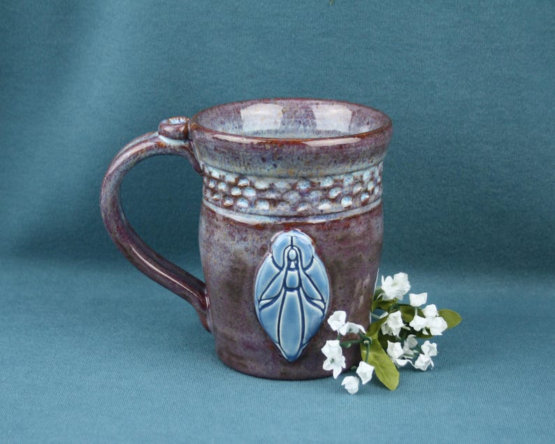 K&ouml;nnte beinhalten: Ein Keramikbecher mit gesprenkelter violetter und blauer Glasur, der ein strukturiertes Band am Rand und ein erhabenes, hellblaues ovales Emblem mit einem stilisierten Insekt aufweist. Ein kleines B&uuml;ndel wei&szlig;er Kunstblumen liegt am Fu&szlig; des Bechers.
