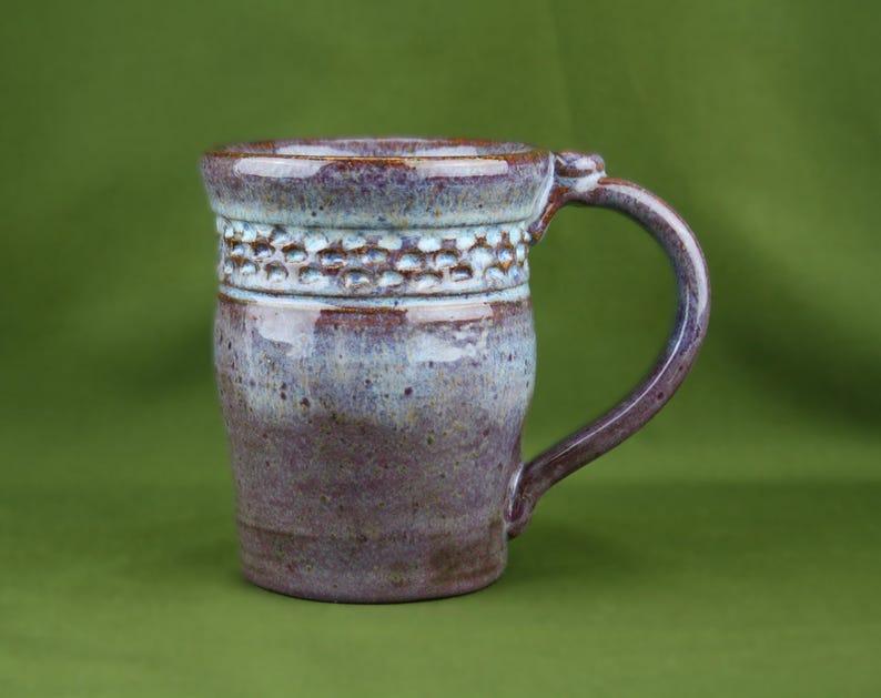 K&ouml;nnte beinhalten: Ein Keramikbecher mit einer gesprenkelten violetten und hellblauen Glasur. Der obere Bereich weist ein strukturiertes Band mit einem sich wiederholenden Wabenmuster auf. Der Becher hat einen stabilen, geschwungenen Henkel und einen glatten Rand.