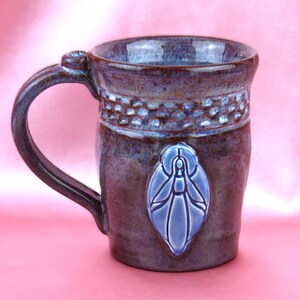 K&ouml;nnte beinhalten: Ein Keramikbecher mit einer gl&auml;nzenden, violett und braun gesprenkelten Glasur. Der Becher hat ein strukturiertes Band am Rand und ein erhabenes, hellblaues, mandelf&ouml;rmiges Emblem mit einer stilisierten Figur auf der Vorderseite.