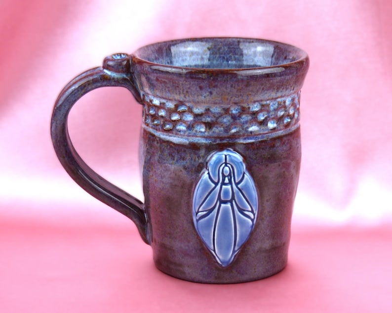K&ouml;nnte beinhalten: Ein Keramikbecher mit einer gl&auml;nzenden, violett und braun gesprenkelten Glasur. Der Becher hat ein strukturiertes Band am Rand und ein erhabenes, hellblaues, mandelf&ouml;rmiges Emblem mit einer stilisierten Figur auf der Vorderseite.