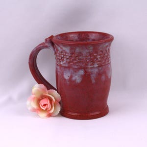 May include: A handmade, red ceramic mug with a textured band near the rim and a large handle. The mug is speckled with darker red and white accents. A pink and cream-colored artificial rose rests near the base of the mug.
