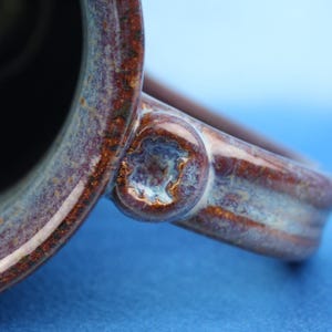K&ouml;nnte beinhalten: Ein Keramikbecher mit einer tiefbraunen und blauen gesprenkelten Glasur mit gl&auml;nzendem Finish. Der Henkel ist mit einem kreisf&ouml;rmigen, strukturierten dekorativen Akzent am Verbindungspunkt an der Seite des Bechers befestigt und zeigt einen dunklen Innenraum.