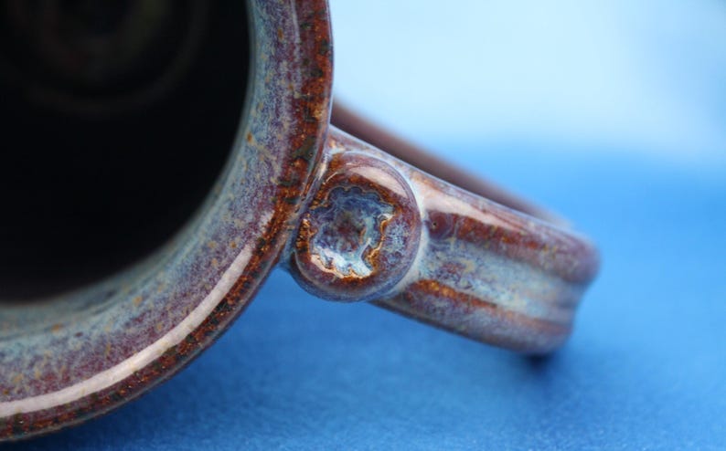 K&ouml;nnte beinhalten: Ein Keramikbecher mit einer tiefbraunen und blauen gesprenkelten Glasur mit gl&auml;nzendem Finish. Der Henkel ist mit einem kreisf&ouml;rmigen, strukturierten dekorativen Akzent am Verbindungspunkt an der Seite des Bechers befestigt und zeigt einen dunklen Innenraum.