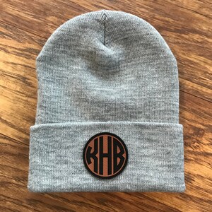 May include: A gray knit beanie with a brown leather patch featuring the initials "RHB" in a circular design. The hat is folded at the brim and sits on a wooden surface.