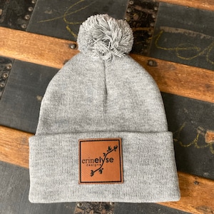 May include: Gray beanie with a pom pom on top and a brown leather patch with the text "erinelyse designs" in black.