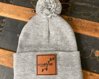 May include: Gray beanie with a pom pom on top and a brown leather patch with the text "erinelyse designs" in black.