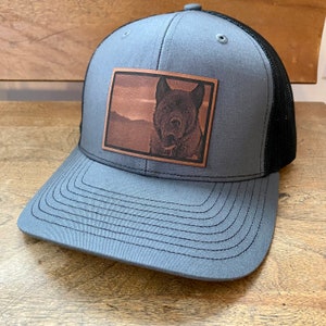 May include: A gray and black trucker hat with a brown leather patch featuring a dog's portrait.