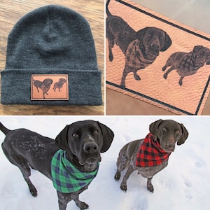 May include: A gray beanie with a brown leather patch featuring two black and brown dogs. The beanie is on a wooden surface. Two black and brown dogs are wearing bandanas and standing on snow.