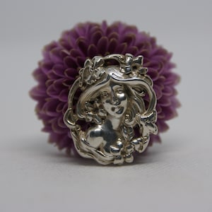 May include: A silver pendant featuring a woman's face surrounded by floral details. The pendant is set against a background of a purple flower.