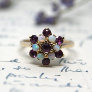 Könnte beinhalten: Ein goldener Ring mit einem blütenförmigen Design, das mit violetten und weißen Edelsteinen besetzt ist. Der Ring liegt auf einer weißen Oberfläche mit handschriftlichem Text.