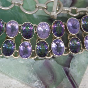 Peut inclure: Un bracelet en argent avec deux rang&eacute;es de pierres pr&eacute;cieuses ovales violettes iris&eacute;es. Le bracelet est dot&eacute; d'un fermoir &agrave; bascule et est pr&eacute;sent&eacute; sur une surface verte et blanche. Les pierres ont diff&eacute;rentes nuances de violet.