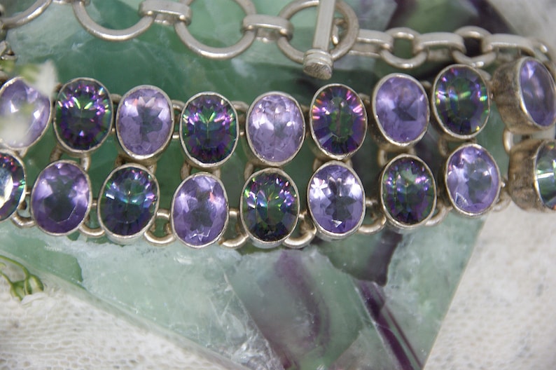 Peut inclure: Un bracelet en argent avec deux rang&eacute;es de pierres pr&eacute;cieuses ovales violettes iris&eacute;es. Le bracelet est dot&eacute; d'un fermoir &agrave; bascule et est pr&eacute;sent&eacute; sur une surface verte et blanche. Les pierres ont diff&eacute;rentes nuances de violet.