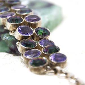 Peut inclure: Gros plan sur un bracelet en argent orn&eacute; de pierres pr&eacute;cieuses ovales violettes iridescentes. Le bracelet est pos&eacute; sur un fond flou vert et blanc. Les pierres ont un &eacute;clat m&eacute;tallique.