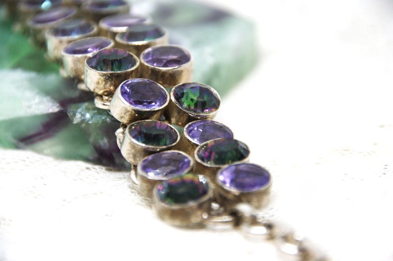 Peut inclure: Gros plan sur un bracelet en argent orn&eacute; de pierres pr&eacute;cieuses ovales violettes iridescentes. Le bracelet est pos&eacute; sur un fond flou vert et blanc. Les pierres ont un &eacute;clat m&eacute;tallique.
