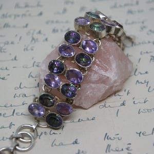Peut inclure: Un bracelet en argent orn&eacute; de plusieurs rang&eacute;es de pierres pr&eacute;cieuses ovales, violettes et iris&eacute;es. Le bracelet est pr&eacute;sent&eacute; sur une pierre rose, sur fond d'&eacute;criture manuscrite. Les pierres pr&eacute;sentent des reflets violets et verts.
