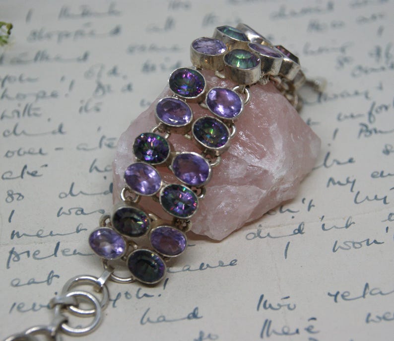 Peut inclure: Un bracelet en argent orn&eacute; de plusieurs rang&eacute;es de pierres pr&eacute;cieuses ovales, violettes et iris&eacute;es. Le bracelet est pr&eacute;sent&eacute; sur une pierre rose, sur fond d'&eacute;criture manuscrite. Les pierres pr&eacute;sentent des reflets violets et verts.