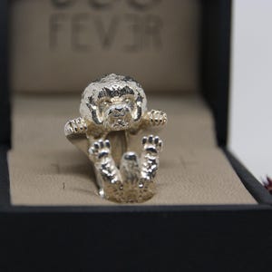 May include: A silver-colored ring featuring a detailed sculpture of a dog. The dog is positioned as if it's lying on its back, with its paws and head raised. The ring is presented in a black box with a beige interior.