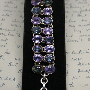 Peut inclure: Un bracelet en argent orn&eacute; de pierres pr&eacute;cieuses ovales violettes iridescentes. Le bracelet est pr&eacute;sent&eacute; sur une surface de velours noir, mettant en valeur le m&eacute;tal poli et les reflets sur les pierres. Le fermoir est une simple bascule.