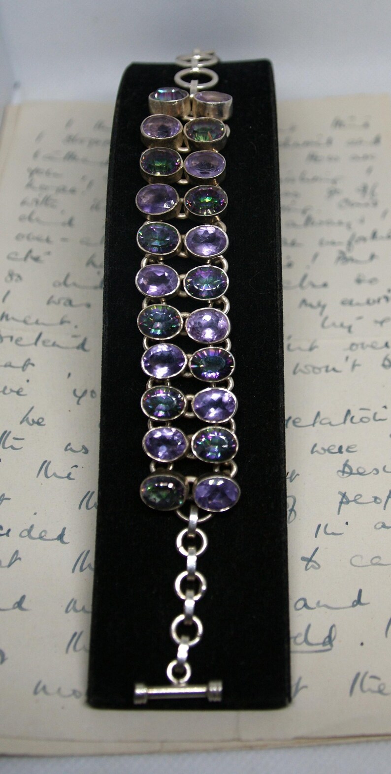 Peut inclure: Un bracelet en argent orn&eacute; de pierres pr&eacute;cieuses ovales violettes iridescentes. Le bracelet est pr&eacute;sent&eacute; sur une surface de velours noir, mettant en valeur le m&eacute;tal poli et les reflets sur les pierres. Le fermoir est une simple bascule.