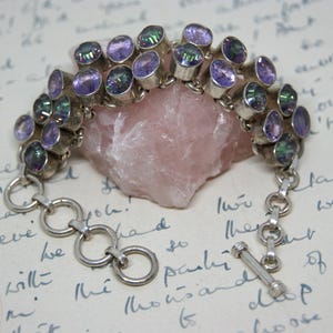 Peut inclure: Un bracelet en argent orn&eacute; de pierres pr&eacute;cieuses ovales, iris&eacute;es et violettes. Le bracelet est compos&eacute; de plusieurs rang&eacute;es de pierres, reli&eacute;es par des maillons en argent et un fermoir &agrave; bascule. Les pierres sont serties dans du m&eacute;tal argent&eacute;.
