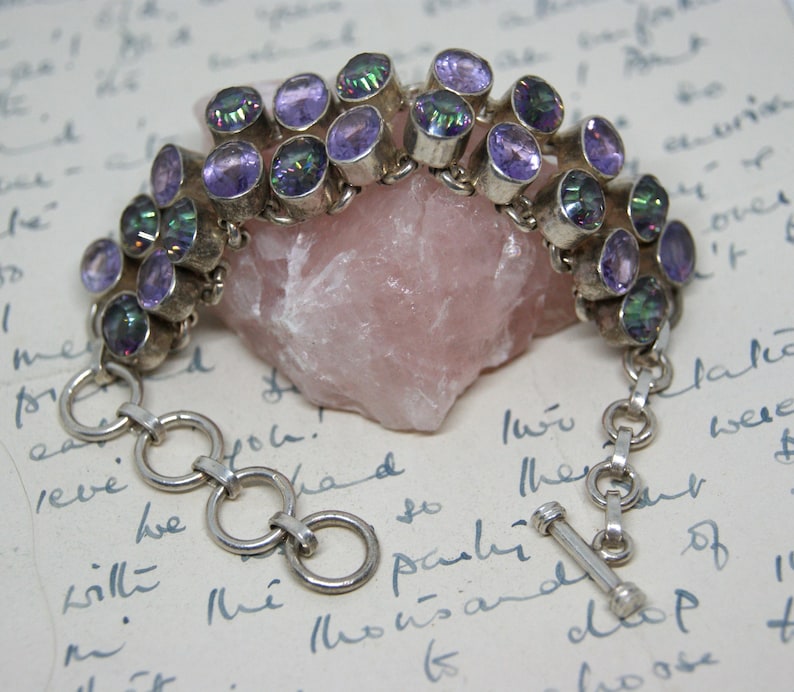 Peut inclure: Un bracelet en argent orn&eacute; de pierres pr&eacute;cieuses ovales, iris&eacute;es et violettes. Le bracelet est compos&eacute; de plusieurs rang&eacute;es de pierres, reli&eacute;es par des maillons en argent et un fermoir &agrave; bascule. Les pierres sont serties dans du m&eacute;tal argent&eacute;.