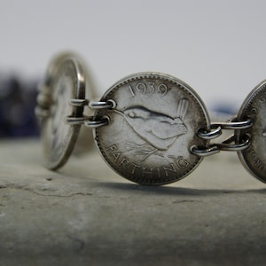 May include: A silver bracelet crafted from linked vintage Farthing coins. The coins display a bird and the year 1939. The bracelet is shown on a textured, grey surface.