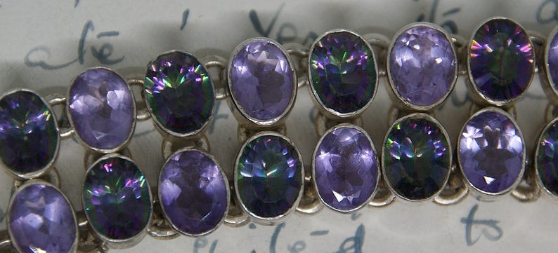 Peut inclure: Un bracelet en argent orn&eacute; de pierres pr&eacute;cieuses ovales dans des tons de violet et de vert iris&eacute;. Les pierres sont serties dans un design de cha&icirc;ne. Le bracelet est un bijou.