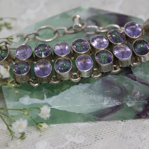 Peut inclure: Un bracelet en argent orn&eacute; de pierres pr&eacute;cieuses ovales, iris&eacute;es, violettes et vertes. Le bracelet est pos&eacute; sur une pierre verte et violette. De petites fleurs blanches sont au premier plan.
