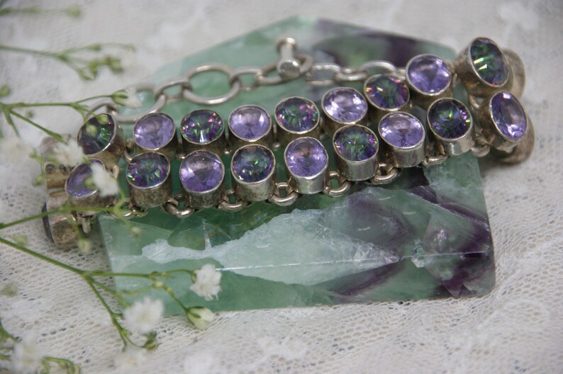 Peut inclure: Un bracelet en argent orn&eacute; de pierres pr&eacute;cieuses ovales, iris&eacute;es, violettes et vertes. Le bracelet est pos&eacute; sur une pierre verte et violette. De petites fleurs blanches sont au premier plan.