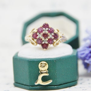 May include: A gold ring with a cluster of red ruby stones and smaller white diamonds set in a square shape. The ring is in a green ring box.