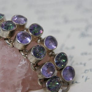 Peut inclure: Un bracelet en argent orn&eacute; de plusieurs pierres pr&eacute;cieuses ovales, violettes et iris&eacute;es. Le bracelet repose sur une pierre rose clair &agrave; texture rugueuse. L'arri&egrave;re-plan est un document manuscrit flou.