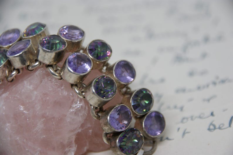 Peut inclure: Un bracelet en argent orn&eacute; de plusieurs pierres pr&eacute;cieuses ovales, violettes et iris&eacute;es. Le bracelet repose sur une pierre rose clair &agrave; texture rugueuse. L'arri&egrave;re-plan est un document manuscrit flou.