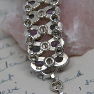 Peut inclure: Gros plan sur un bracelet en argent avec des maillons ovales contenant chacun une petite pierre pr&eacute;cieuse violet fonc&eacute;. Le bracelet est reli&eacute; par de petits maillons circulaires. Le fond comprend une pierre rose et une &eacute;criture manuscrite.