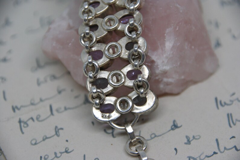 Peut inclure: Gros plan sur un bracelet en argent avec des maillons ovales contenant chacun une petite pierre pr&eacute;cieuse violet fonc&eacute;. Le bracelet est reli&eacute; par de petits maillons circulaires. Le fond comprend une pierre rose et une &eacute;criture manuscrite.